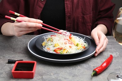 Photo of Stir-fry. Woman with chopsticks eating tasty rice noodles with meat and vegetables at grey textured table, closeup