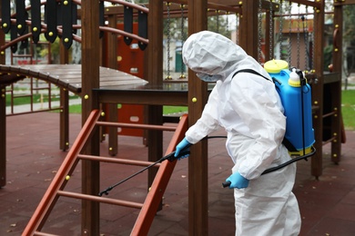 Photo of Woman wearing chemical protective suit with disinfectant sprayer on playground. Preventive measure during coronavirus pandemic