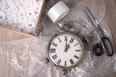 Vintage alarm clock with bubble wrap, scissors and adhesive tape on wooden table, flat lay