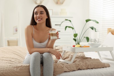 Happy young woman having breakfast near white tray on bed at home