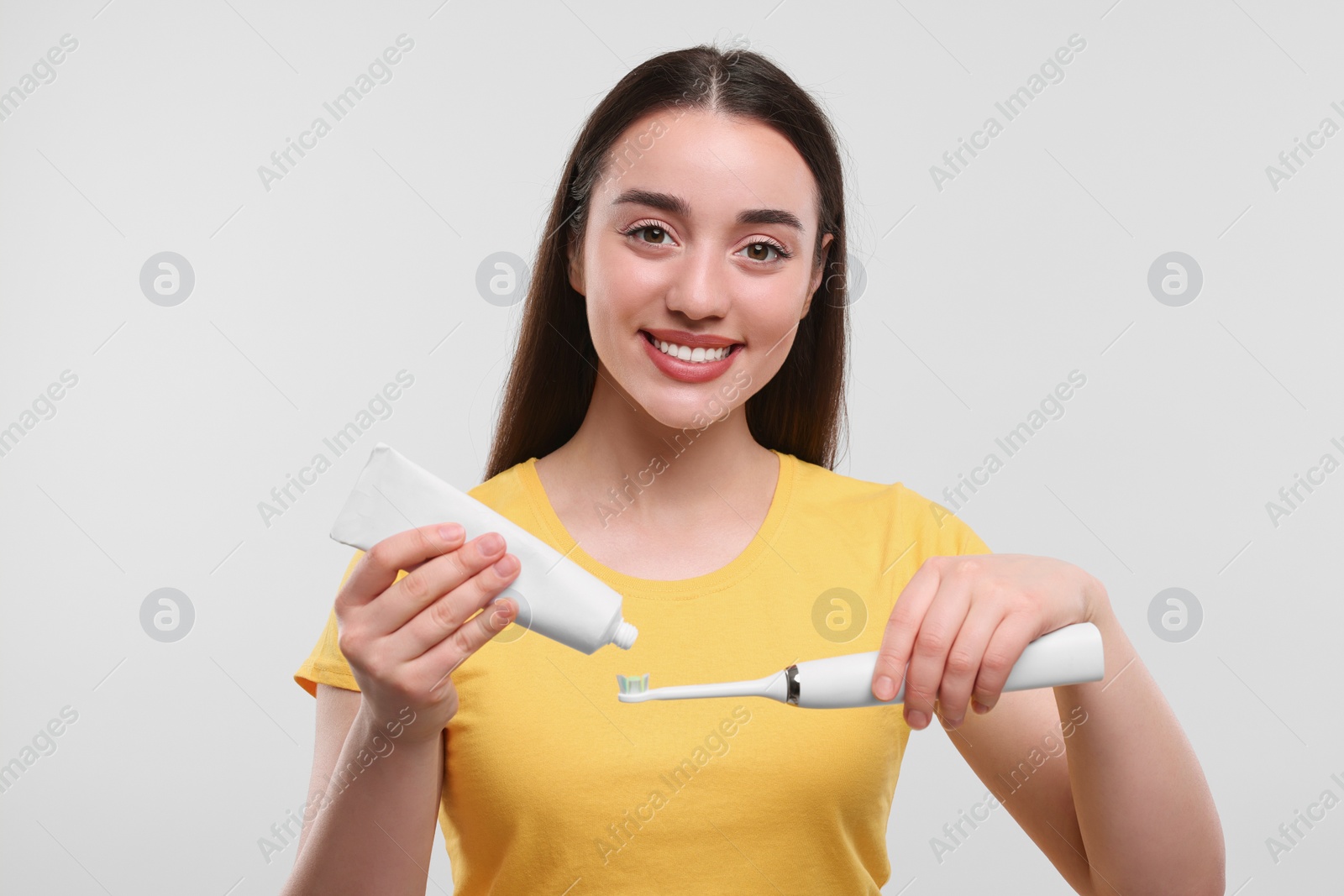 Photo of Happy young woman squeezing toothpaste from tube onto electric toothbrush on white background