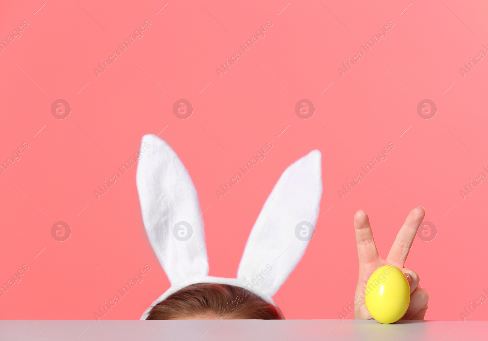 Photo of Little girl wearing bunny ears headband  and playing with Easter egg at table against color background