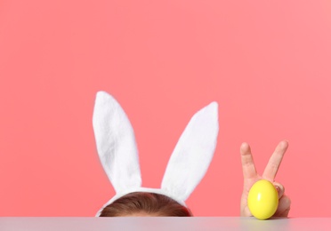 Little girl wearing bunny ears headband  and playing with Easter egg at table against color background