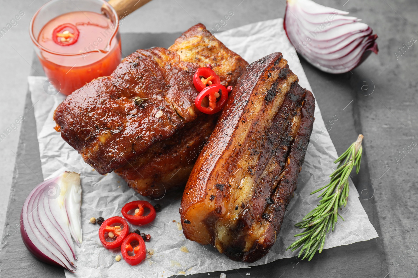 Photo of Pieces of baked pork belly served with sauce, rosemary and chili pepper on grey table