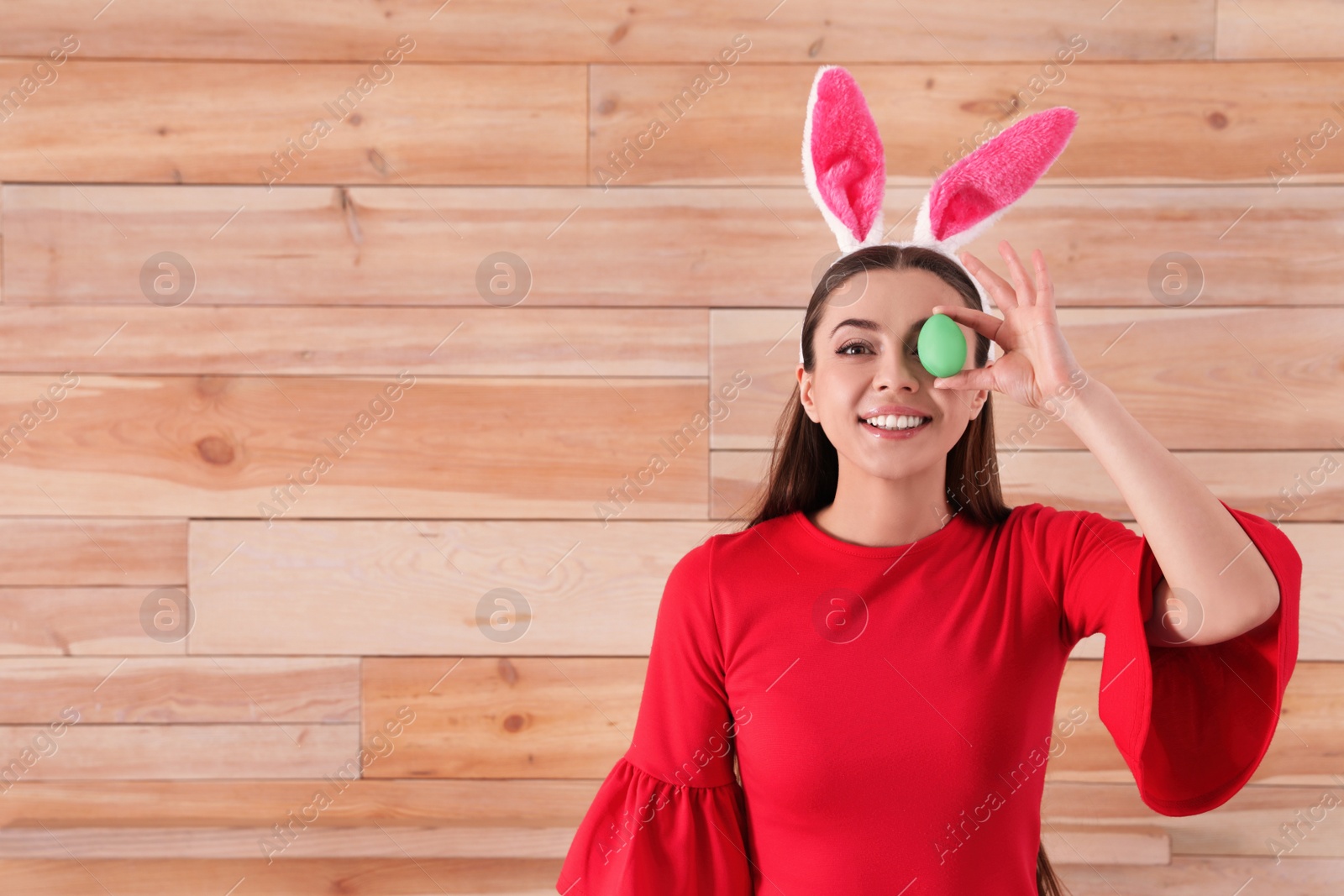 Photo of Beautiful woman in bunny ears headband holding Easter egg near eye against wooden background, space for text