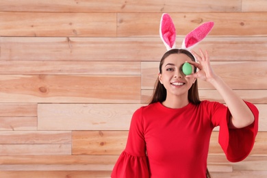 Photo of Beautiful woman in bunny ears headband holding Easter egg near eye against wooden background, space for text