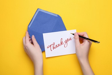 Woman with envelope and card writing phrase Thank You on yellow background, top view