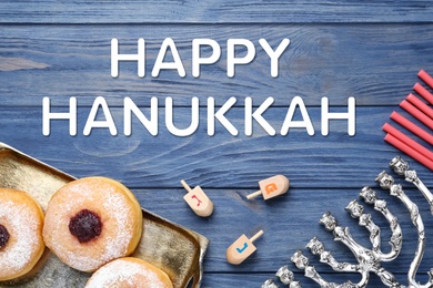 Image of Happy Hanukkah. Flat lay composition with menorah, red candles, dreidels and sufganiyot on blue wooden table