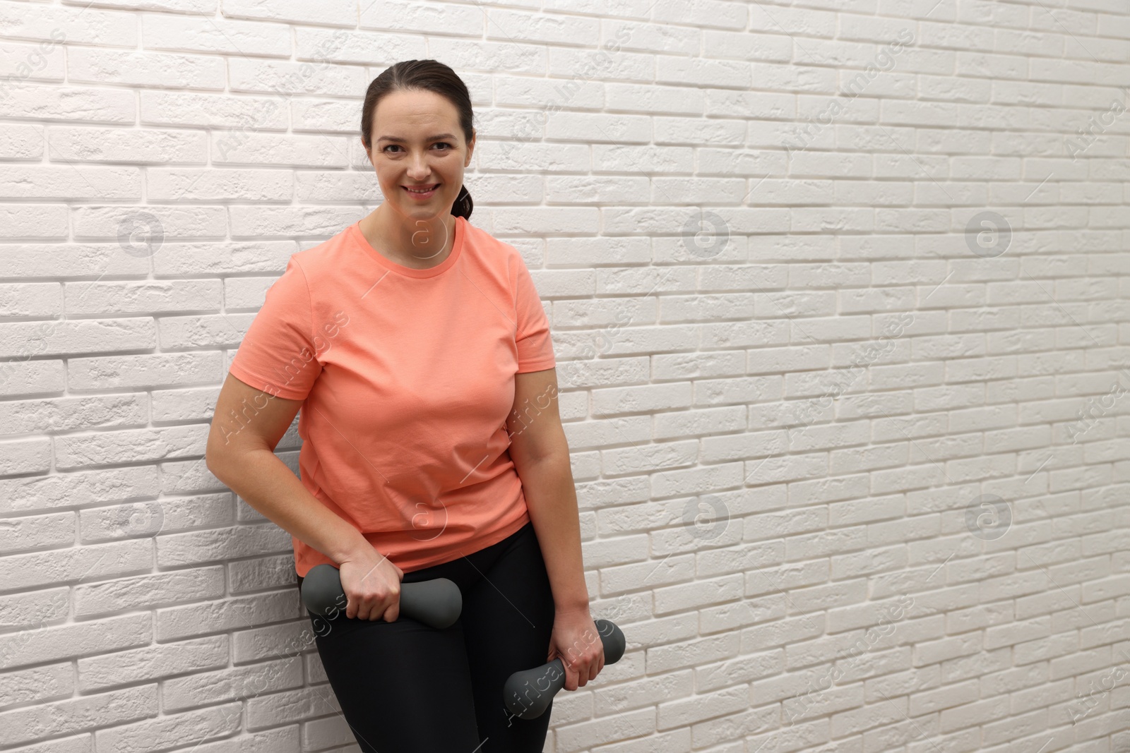 Photo of Happy overweight woman with dumbbells near white brick wall, space for text