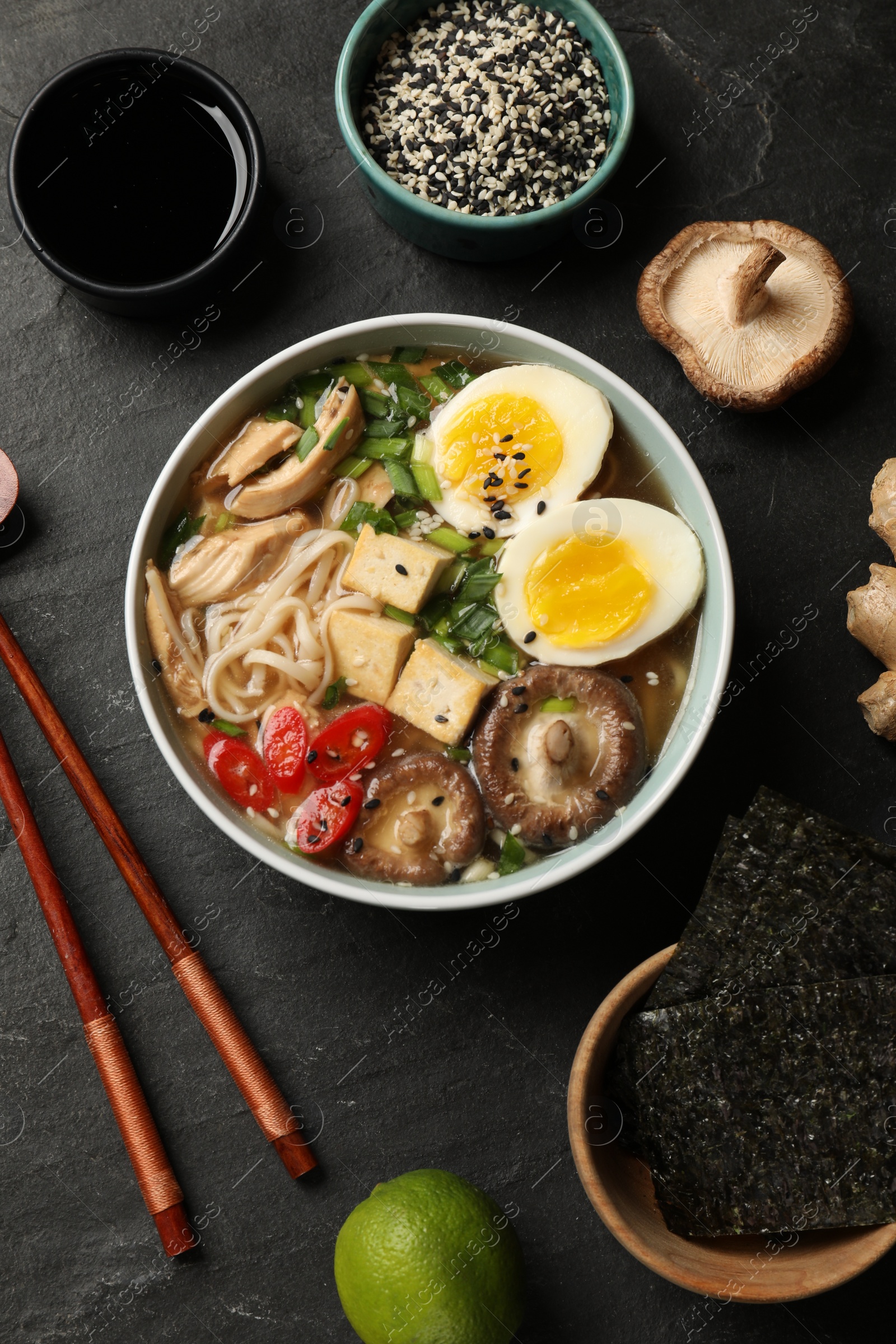 Photo of Bowl of delicious ramen, ingredients and chopsticks on black table, flat lay. Noodle soup