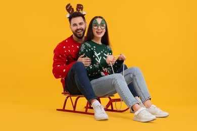Photo of Happy young couple in Christmas sweaters, reindeer headband and party glasses on sled against orange background