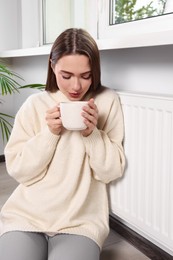 Photo of Woman holding cup with hot drink near heating radiator indoors