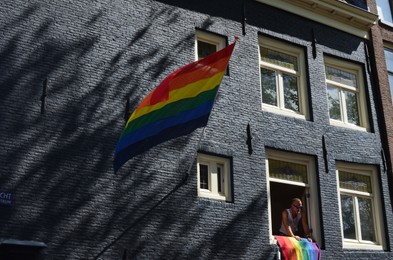 Photo of AMSTERDAM, NETHERLANDS - AUGUST 06, 2022: Building facade with bright LGBT pride flags and man looking out of window at parade