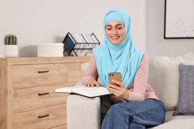 Muslim woman with book using smartphone on couch in room