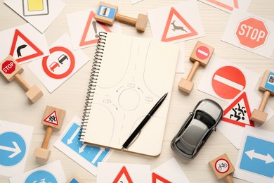 Many different road signs, notebook with sketch of roundabout and toy car on white wooden background, flat lay. Driving school