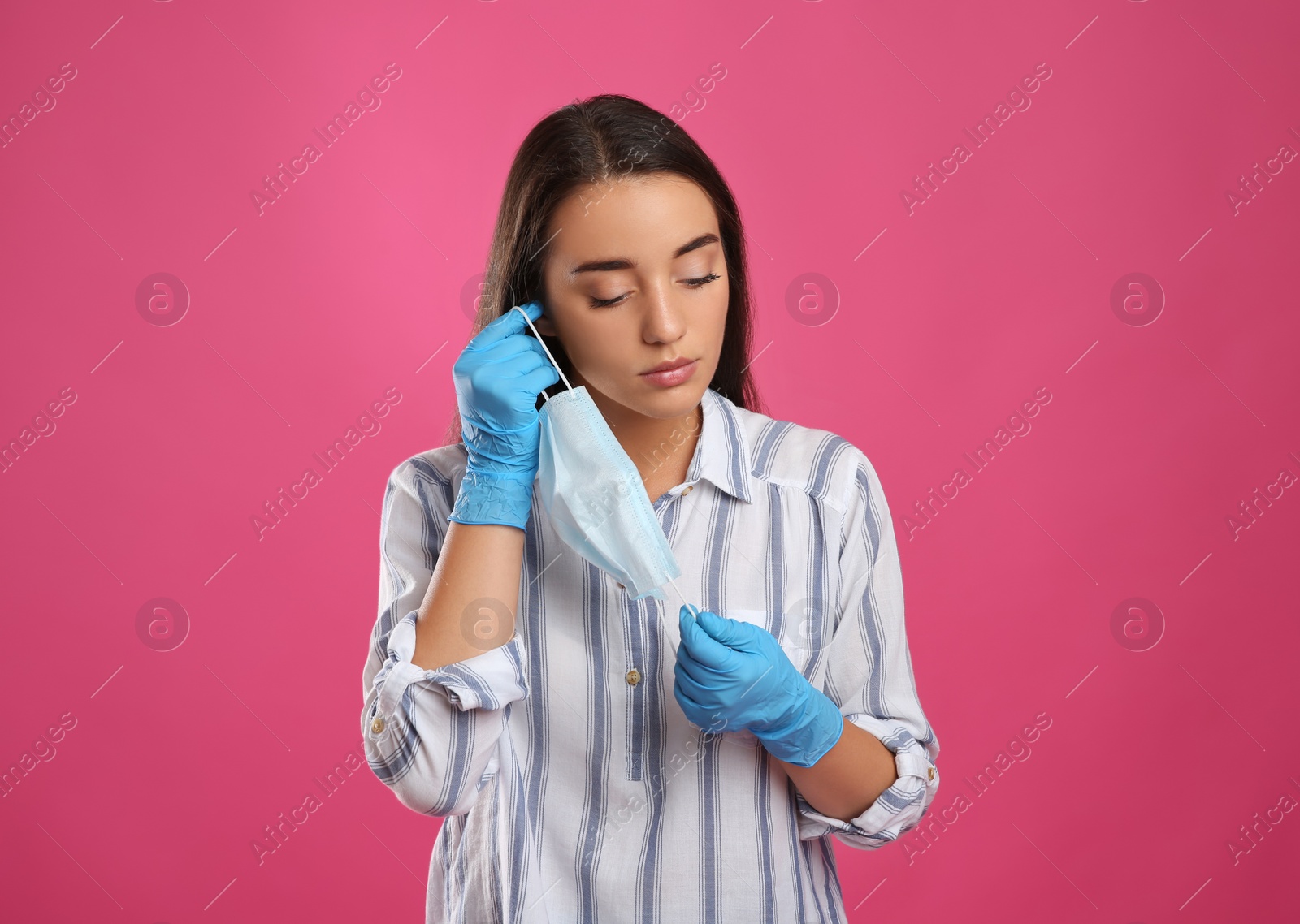 Photo of Woman in medical gloves putting on protective face mask against pink background