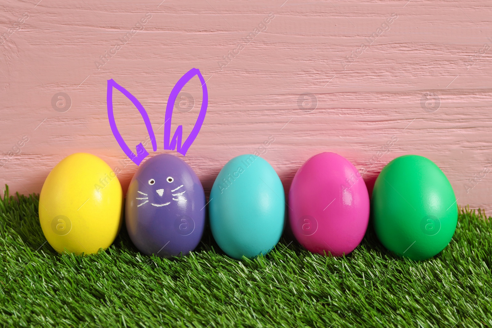 Image of Violet eggs with drawn face and ears as Easter bunny among others on green grass against pink wooden background