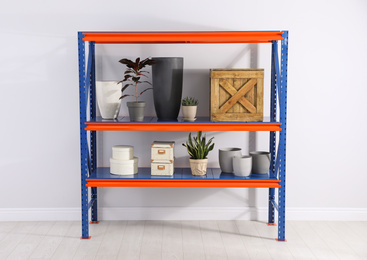 Photo of Metal shelving unit with different household stuff near light wall indoors