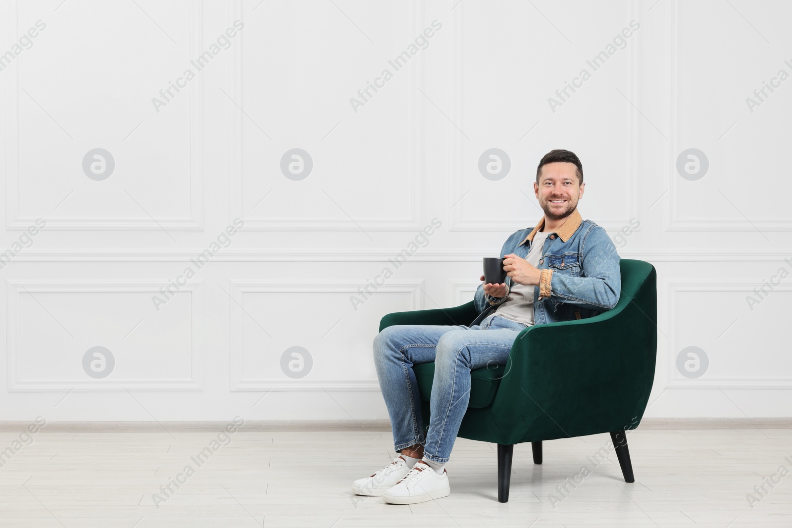 Photo of Happy man with cup of drink sitting in armchair indoors, space for text