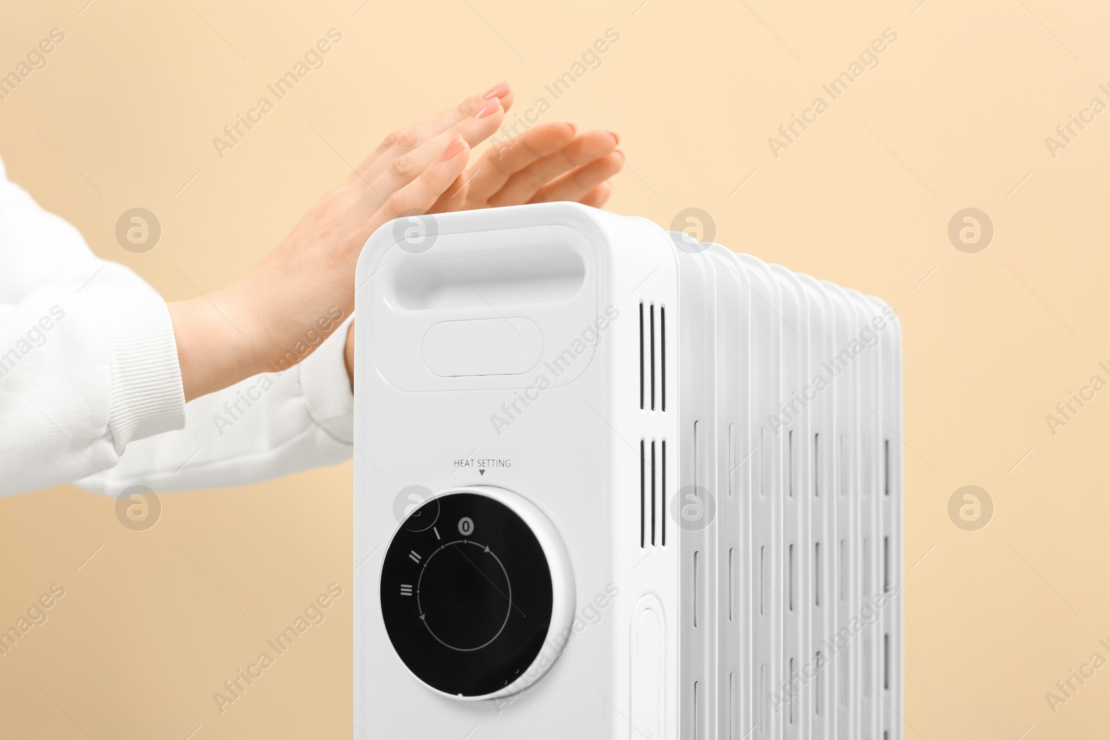 Photo of Young woman warming hands near modern electric heater on beige background, closeup