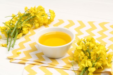 Photo of Rapeseed oil in bowl and beautiful yellow flowers on white table