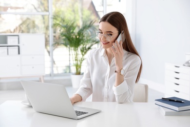 Photo of Young businesswoman talking on phone while using laptop at table in office