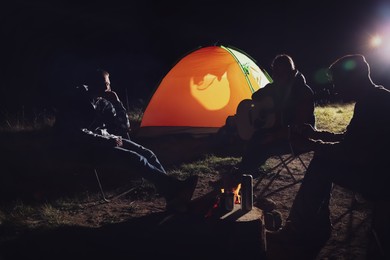 Photo of Group of friends with guitar near bonfire and camping tent outdoors at night