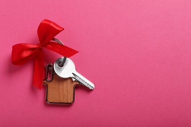 Key with trinket in shape of house and bow on pink background, top view. Space for text. Housewarming party