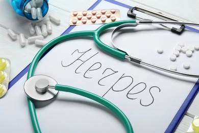 Photo of Clipboard with word Herpes, different pills and stethoscope on white wooden table, closeup