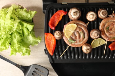 Photo of Electric grill with homemade sausages, vegetables and spatula on wooden table, flat lay