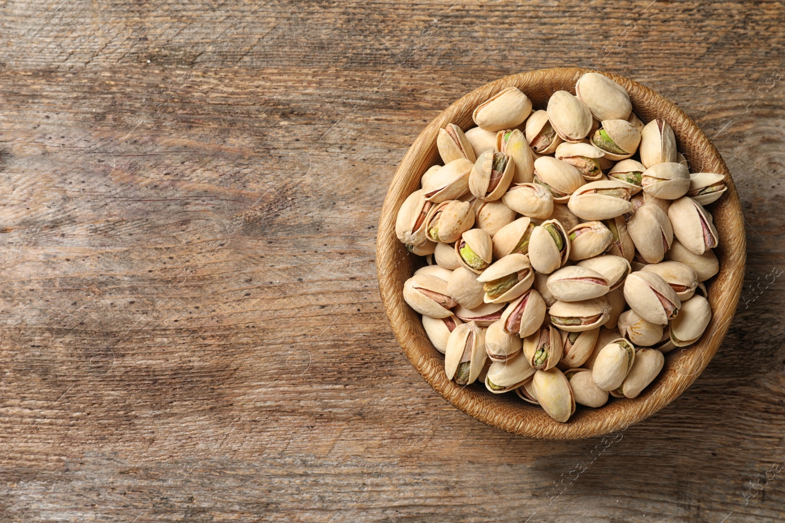 Photo of Organic pistachio nuts in bowl on wooden table, top view. Space for text