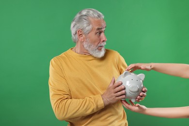 Photo of Woman taking piggy bank from confused senior man on green background. Be careful - fraud