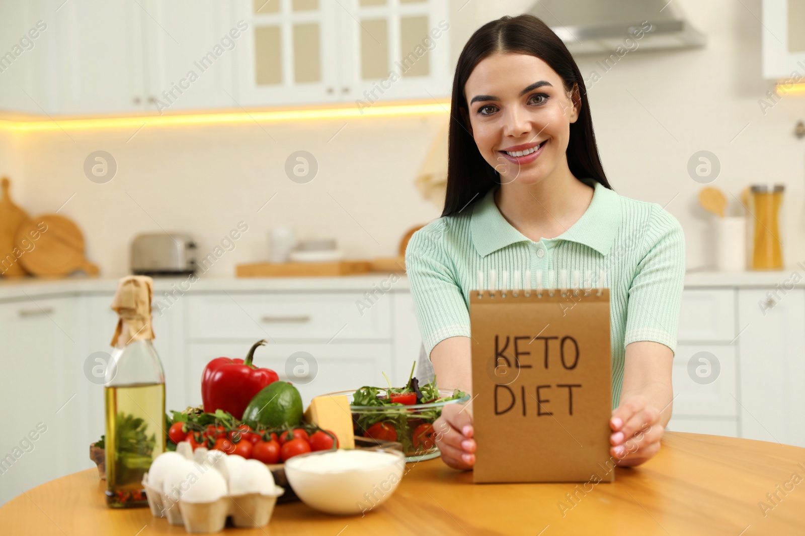 Photo of Happy woman holding notebook with words Keto Diet near different products in kitchen