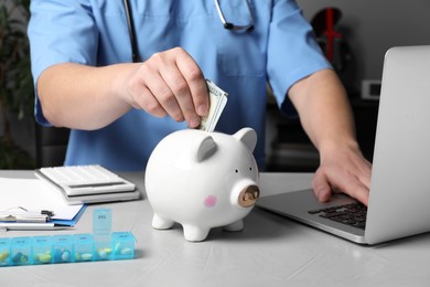 Doctor putting banknote into piggy bank at table in hospital, closeup. Medical insurance