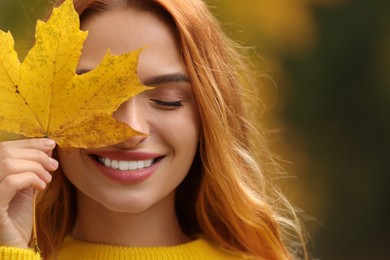 Photo of Smiling woman covering eye with autumn leaf outdoors, closeup. Space for text