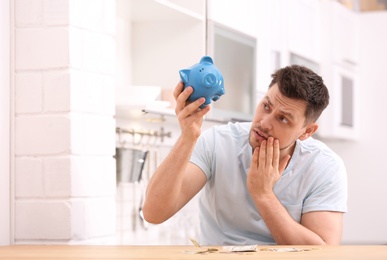 Photo of Sad man with piggy bank and money at home. Space for text