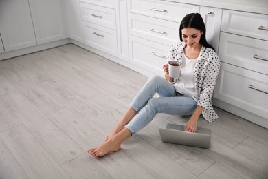 Photo of Happy woman with laptop and cup of drink sitting on warm floor in kitchen, space for text. Heating system