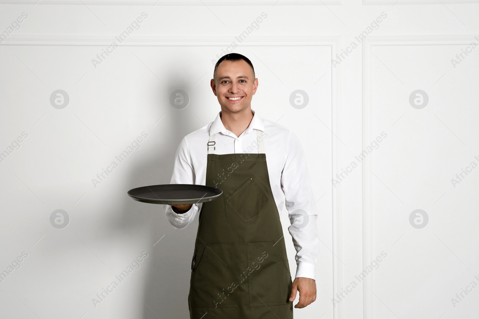 Photo of Portrait of happy young waiter with tray near white wall