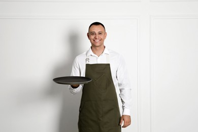 Photo of Portrait of happy young waiter with tray near white wall
