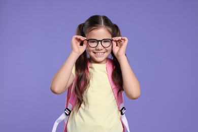 Photo of Cute schoolgirl in glasses with backpack on violet background
