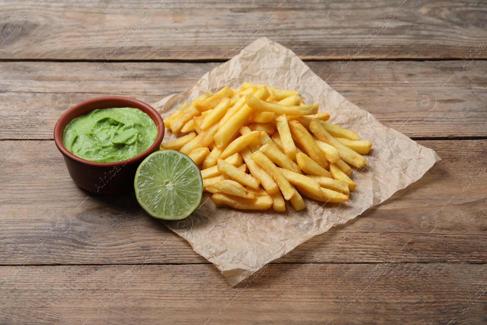 Photo of Parchment with delicious french fries, avocado dip and lime served on wooden table