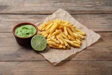 Photo of Parchment with delicious french fries, avocado dip and lime served on wooden table