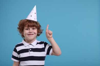 Photo of Cute little boy in party hat pointing at something on light blue background. Space for text