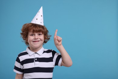 Cute little boy in party hat pointing at something on light blue background. Space for text