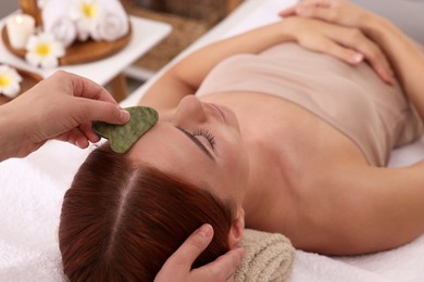 Photo of Young woman receiving facial massage with jade gua sha tool in beauty salon, closeup