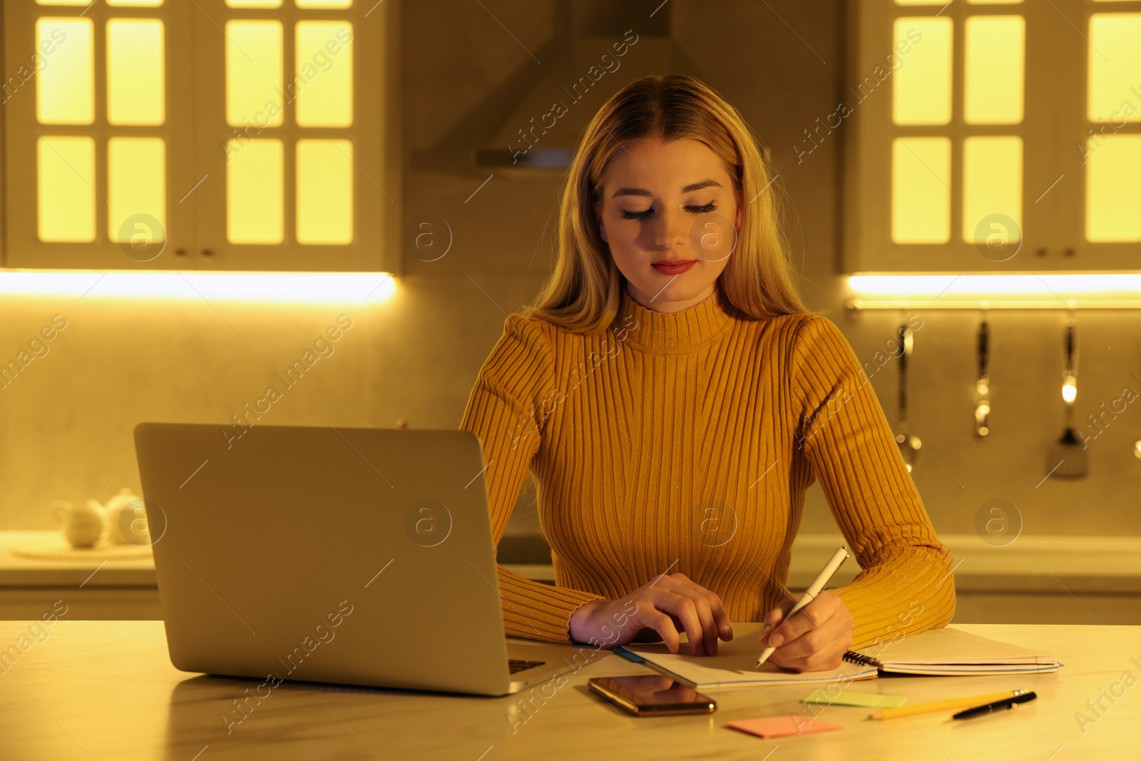 Photo of Home workplace. Woman writing in notebook near laptop at marble desk in kitchen