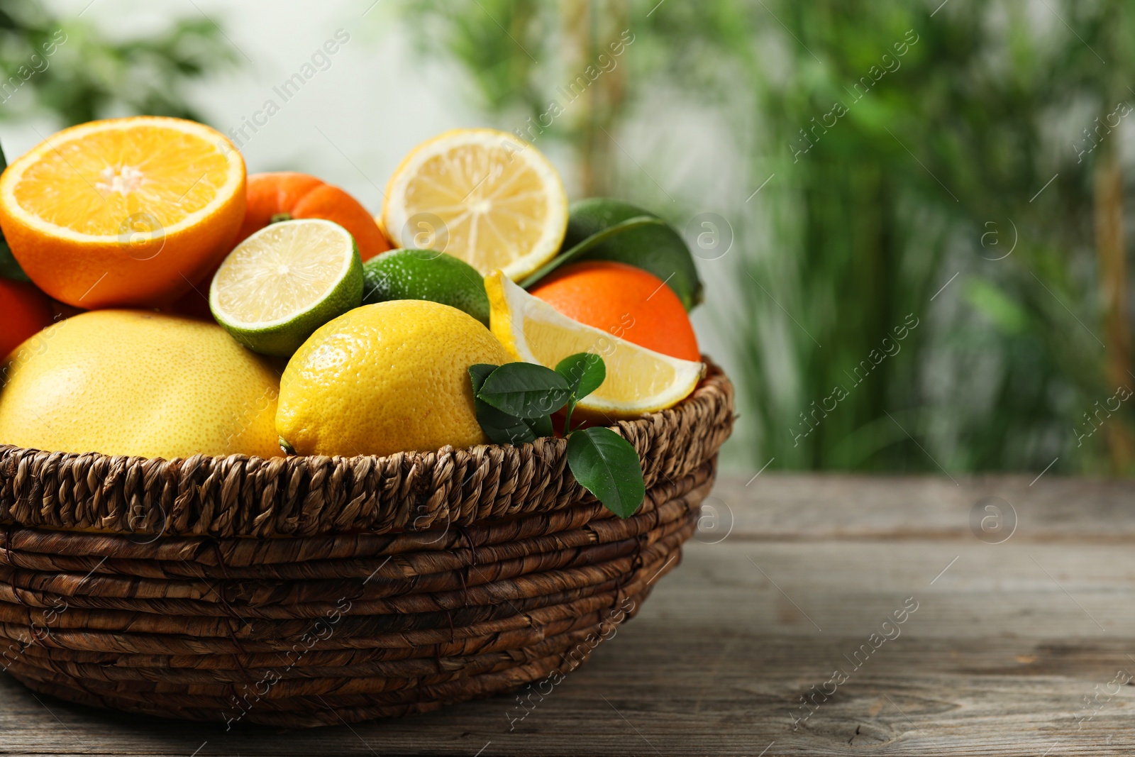 Photo of Different fresh citrus fruits and leaves in wicker basket on wooden table against blurred background, closeup. Space for text