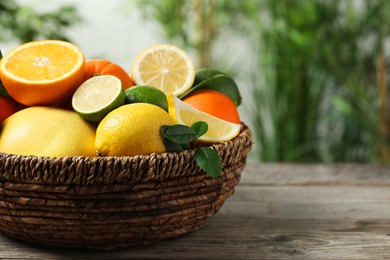 Photo of Different fresh citrus fruits and leaves in wicker basket on wooden table against blurred background, closeup. Space for text