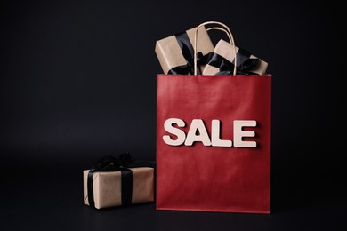 Photo of Red shopping bag with word Sale and gift boxes on dark background. Black Friday