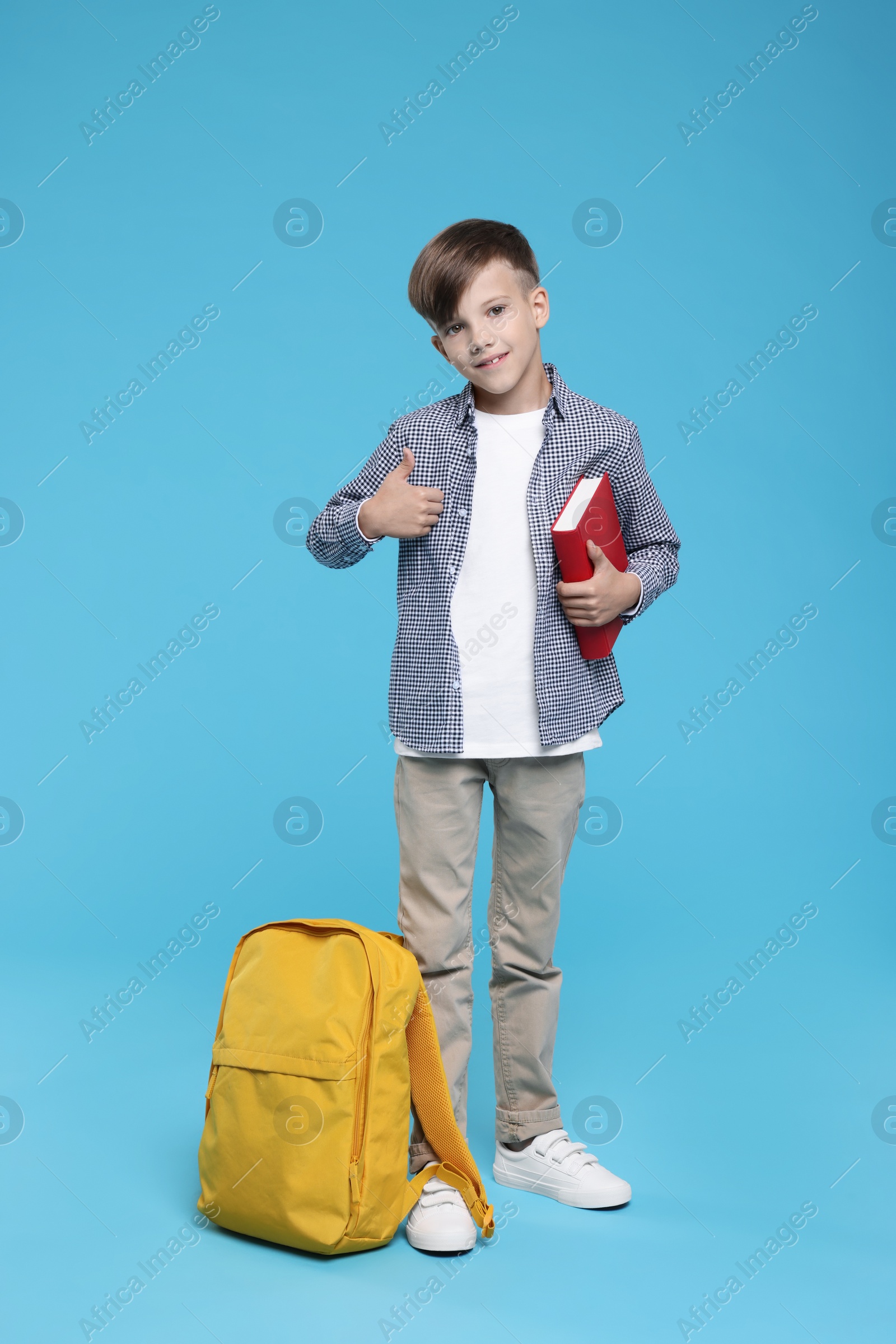 Photo of Cute schoolboy with book showing thumbs up on light blue background